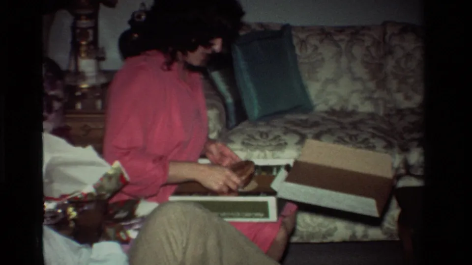 A woman in pink examines an item or wrapping inside an open gift box on her lap, seated beside a man in the 1981 home living room.