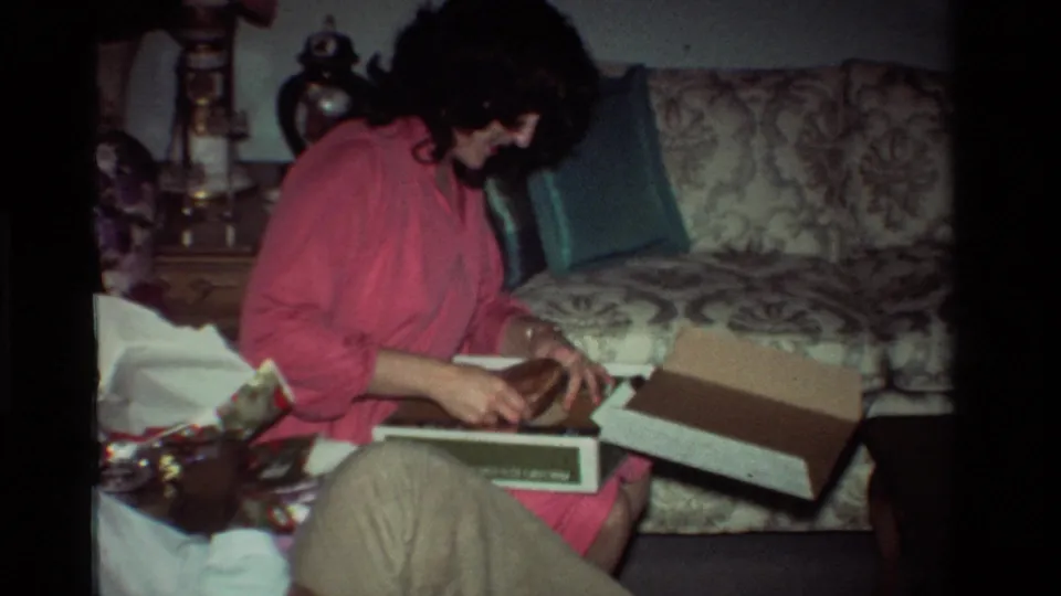 The woman in pink smiles or looks pleased while seated with the open gift box in a 1981 California living room, with the man partly visible beside her.