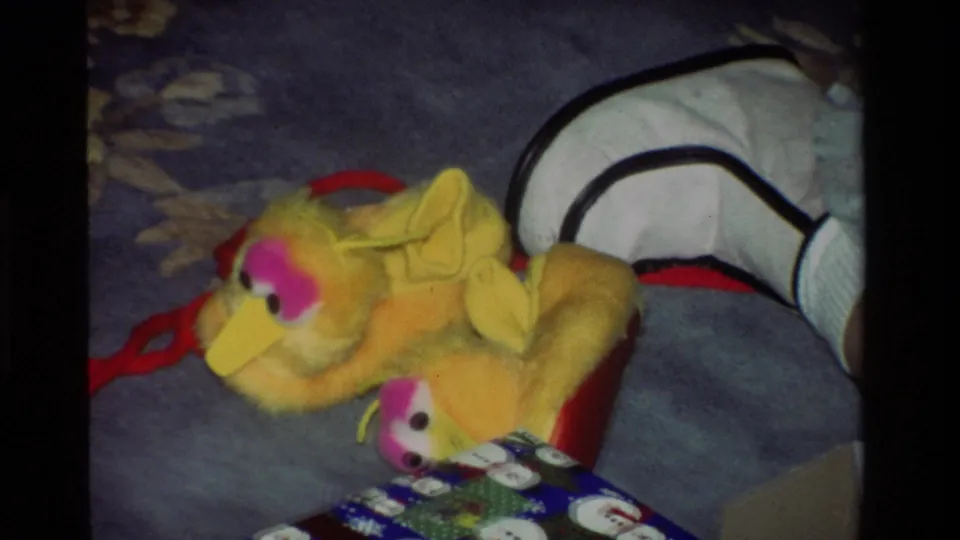 The yellow plush bird toy remains on the blue carpet, with holiday wrapping and a wrapped box visible in the foreground.