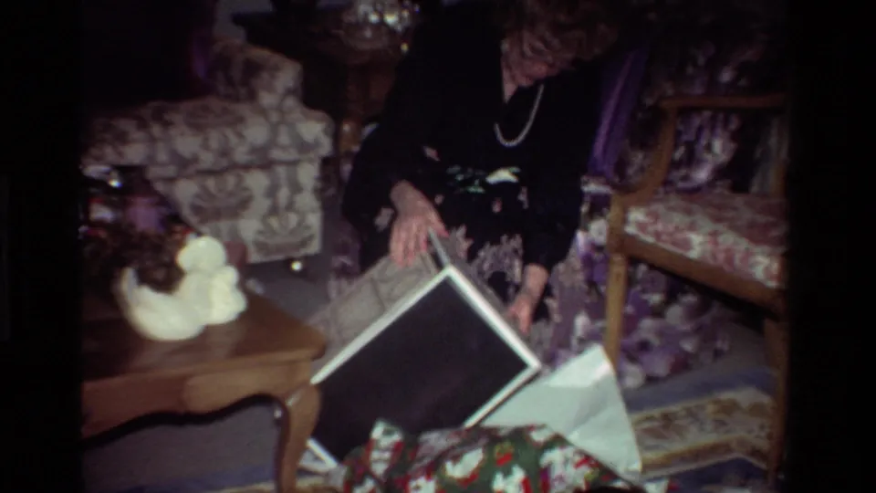A woman in a dark dress and pearl necklace opens or handles a large gift box in a decorated California living room.