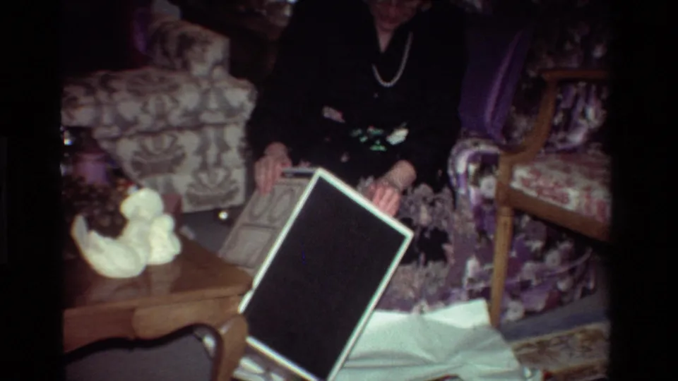 The woman holds an open gift box in the living room, surrounded by patterned chairs, a coffee table, and holiday clutter.