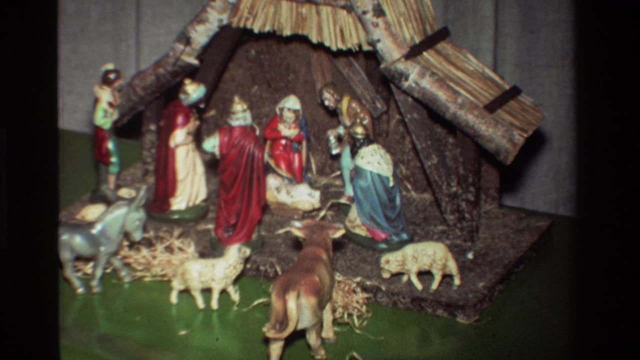 1981 8mm home-movie frame of robed nativity figures around the manger, with sheep, donkey, cow, straw, and a thatched stable on a green base.