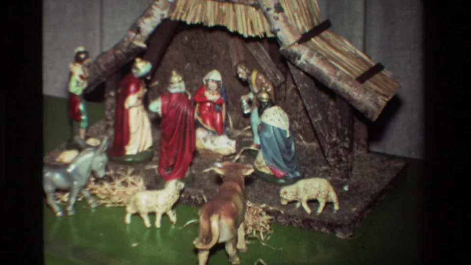 1981 8mm home-movie frame of robed nativity figures around the manger, with sheep, donkey, cow, straw, and a thatched stable on a green base.