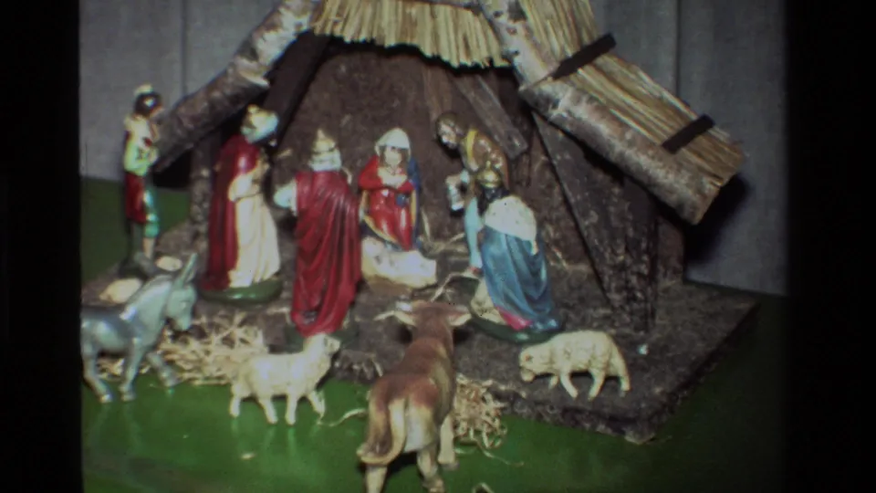Closer 1981 8mm frame of a Christmas nativity arrangement with Mary, Joseph, baby Jesus, visitors, straw, sheep, donkey, cow, and a thatched stable.