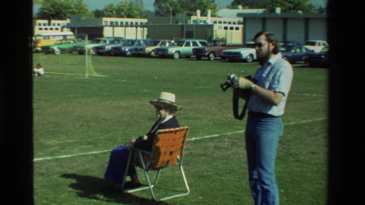 In California 1981 home-movie footage, a bearded man with sunglasses holds a camera beside a seated woman in a hat on a grassy field.