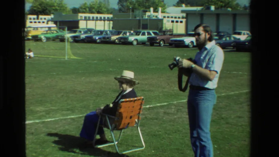 In California 1981 home-movie footage, a bearded man with sunglasses holds a camera beside a seated woman in a hat on a grassy field.