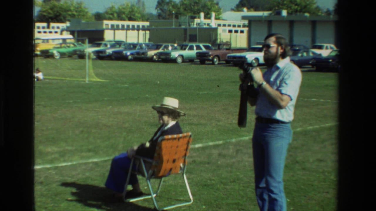 The man raises a still camera toward his eye near a seated woman, with parked cars and a low building behind the grassy field.