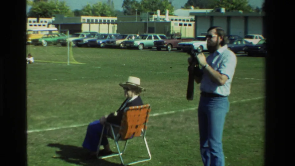 The man raises a still camera toward his eye near a seated woman, with parked cars and a low building behind the grassy field.