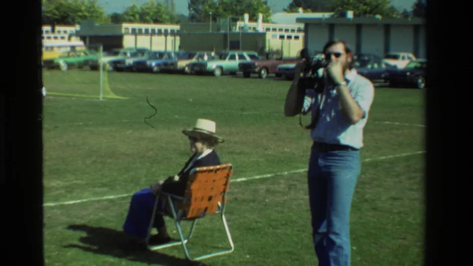 A bearded man in sunglasses aims his camera on a California field in 1981, with a seated woman, cars, and a soccer goal visible.