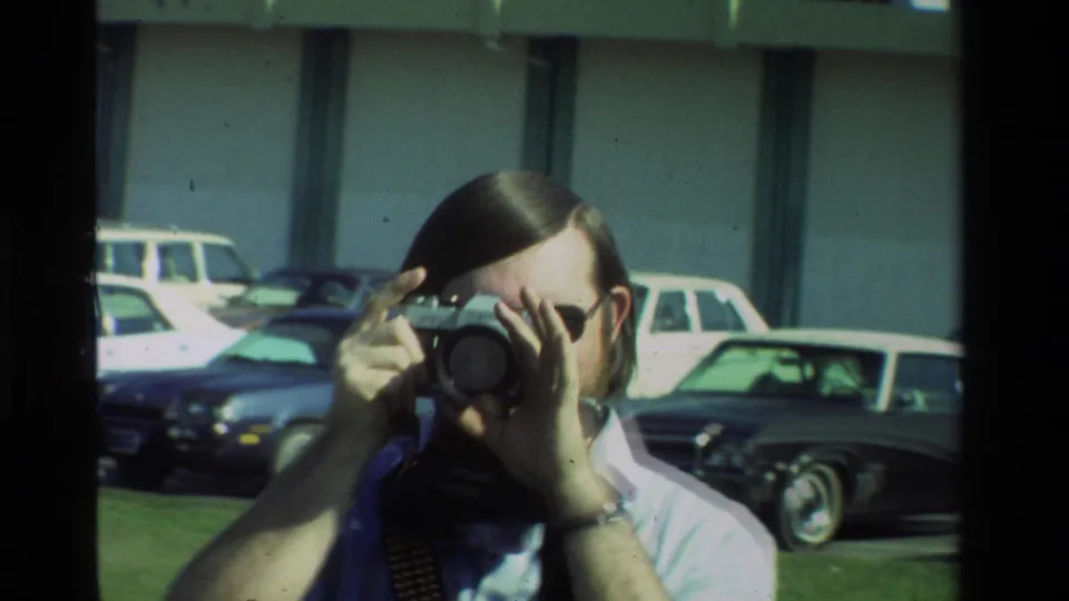 Close-up of the amateur photographer looking through his camera, with parked cars and a building blurred behind him.