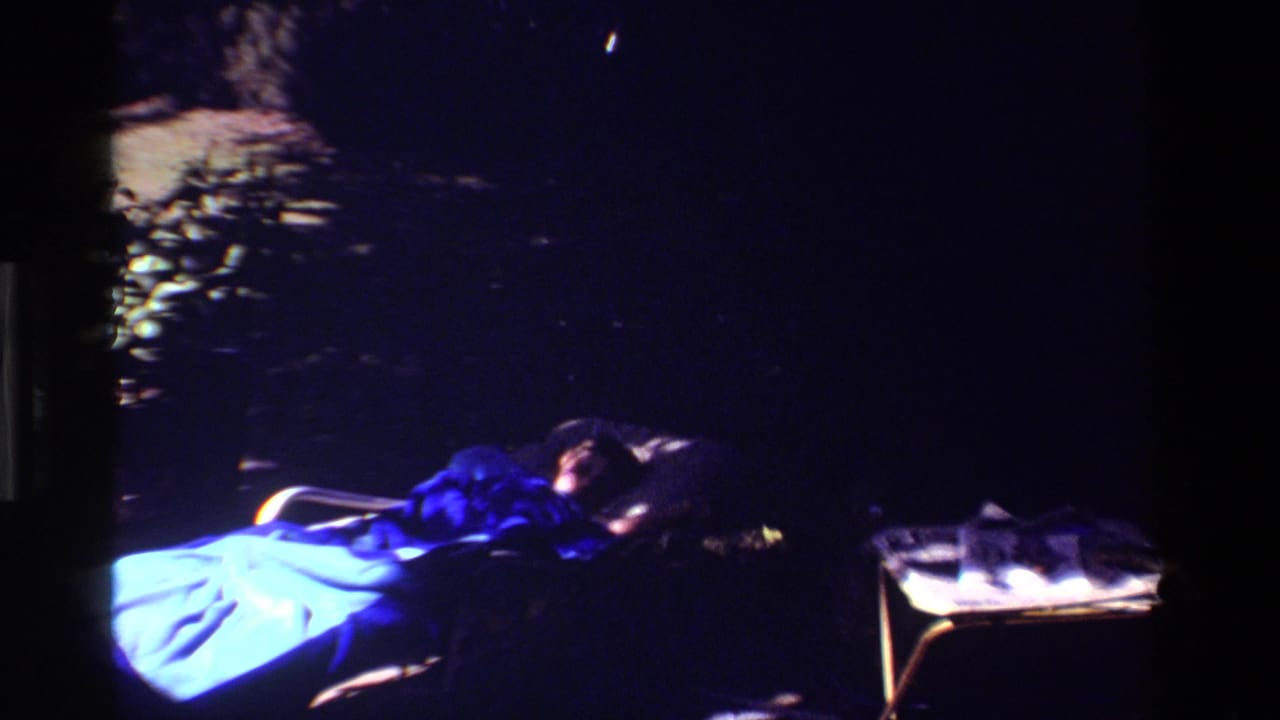 Woman resting on a lawn chair outdoors at night under a blue blanket beside a small table.