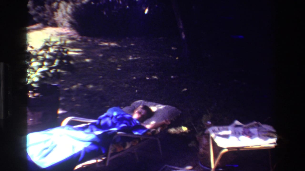 1981 California home-movie frame with a woman lying on a lawn chair at night, lit by cool blue light, with a side table and newspaper pages visible.