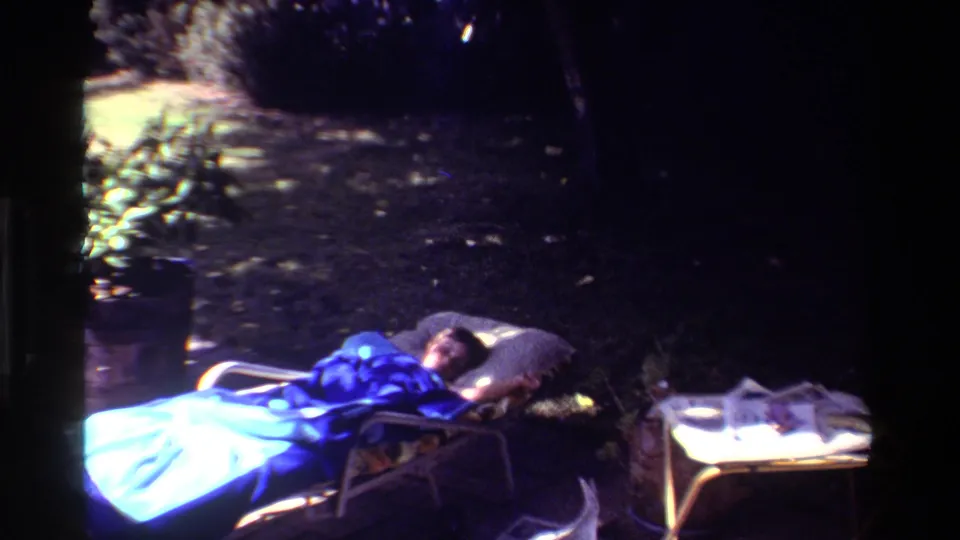 1981 California frame showing a woman resting in a dark yard on a reclining chair, covered by a blue blanket with loose newspaper pages nearby.