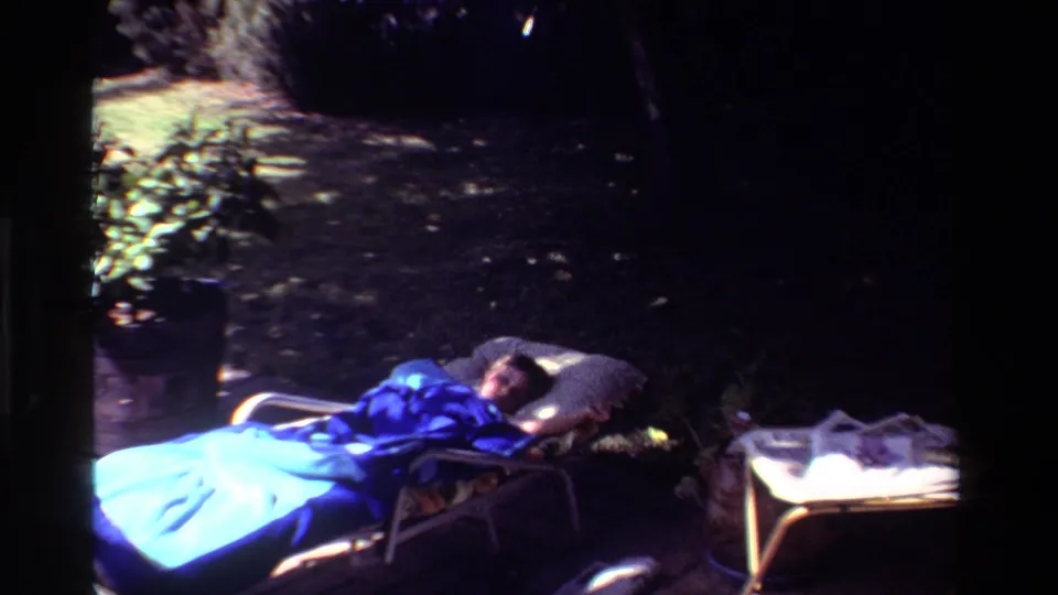 Final 1981 California home-movie frame of a woman reclining at night in a yard, with blue blanket, pillow, side table, and newspaper pages visible.