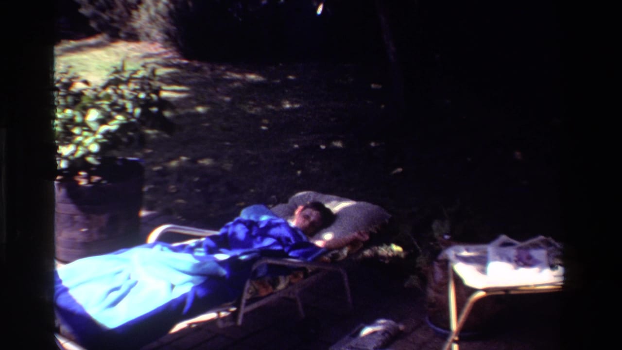 1981 California home-movie frame of a person napping on a chaise lounge with a blue blanket, pillow, side table, and potted plant nearby.