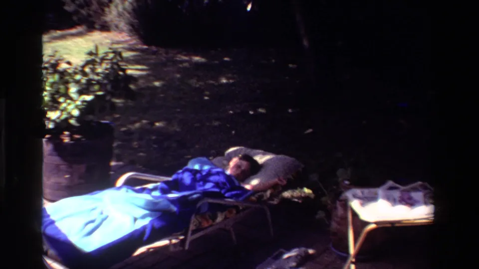 1981 California backyard footage shows a person resting on a chaise lounge, wrapped in a blue blanket beside a small outdoor table.