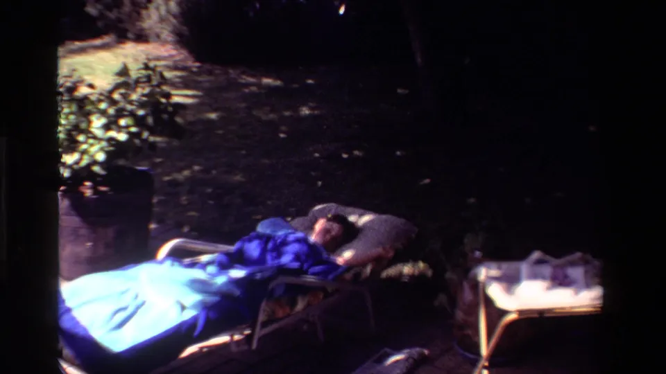 A California 1981 home-movie still shows a person napping outdoors under a blue blanket near a potted plant and small side table.