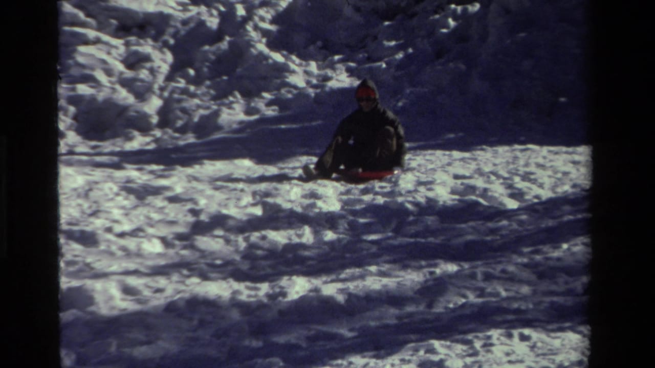 The child continues sledding through a sunlit snowy area, seen from a stationary 8mm home-movie camera.