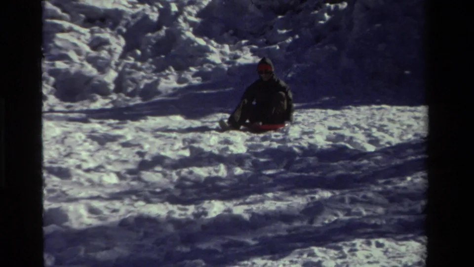 The child continues sledding through a sunlit snowy area, seen from a stationary 8mm home-movie camera.