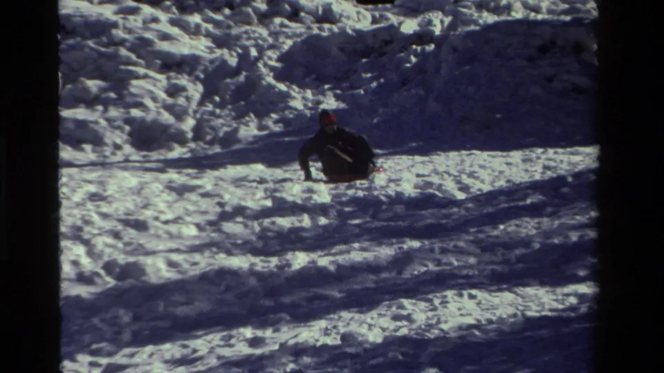 The sledder leans on the red saucer sled near the lower part of the snowy slope, with blue shadows across the snow.