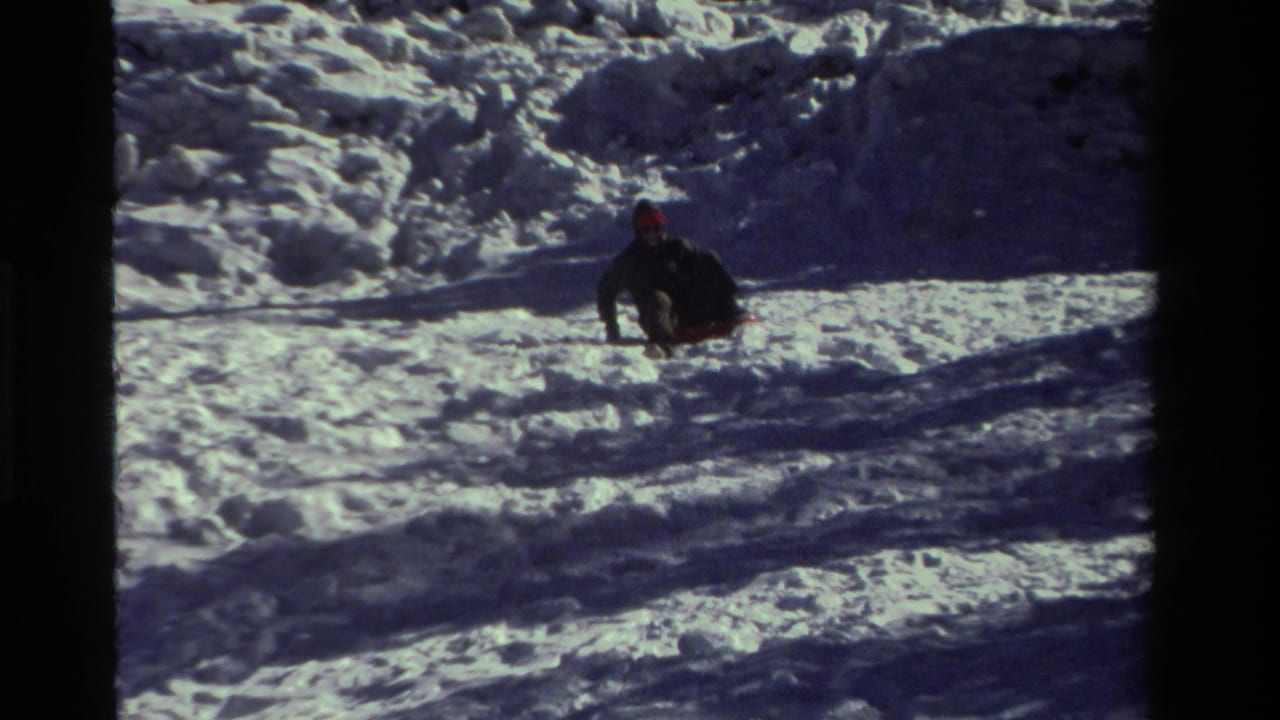 1981 California 8mm frame showing a distant person in dark winter clothing and a red hat seated on a sled amid uneven snow.