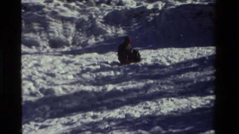 1981 California home-movie frame with a distant sledder in dark clothing crossing a rutted snowy slope.