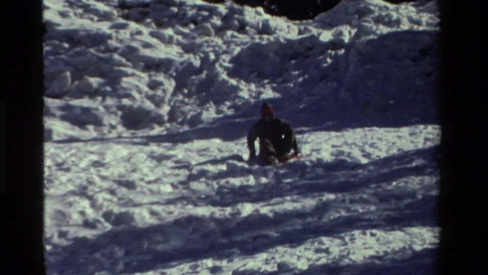 Distant 1981 California 8mm view of a sledder sitting low on a snowy slope with dark clothing and a red hat visible.