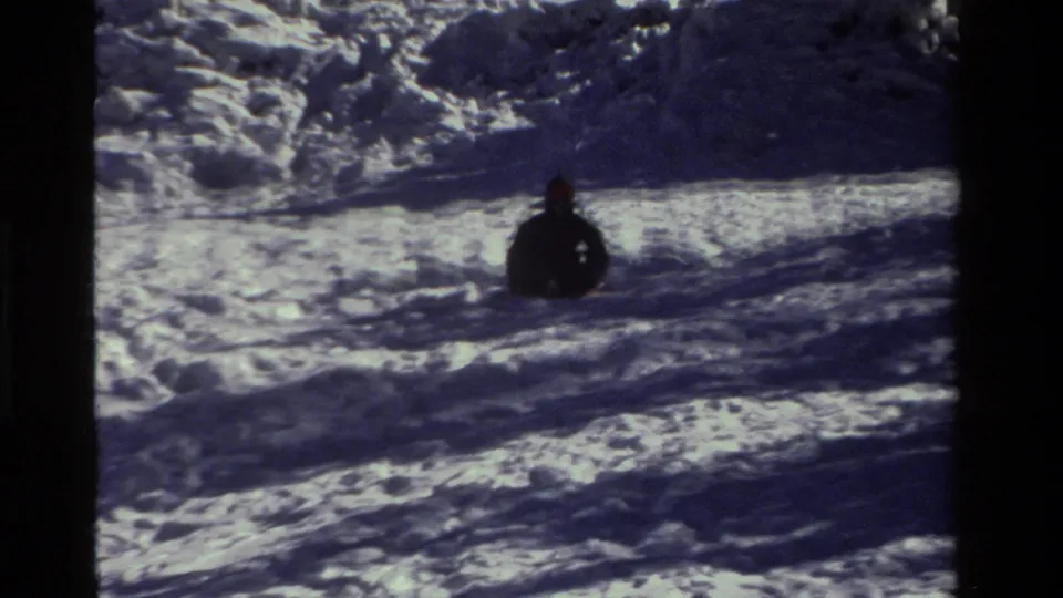 1981 California frame showing the sledder from behind, seated in dark clothing on a snowy hill with strong shadow bands.