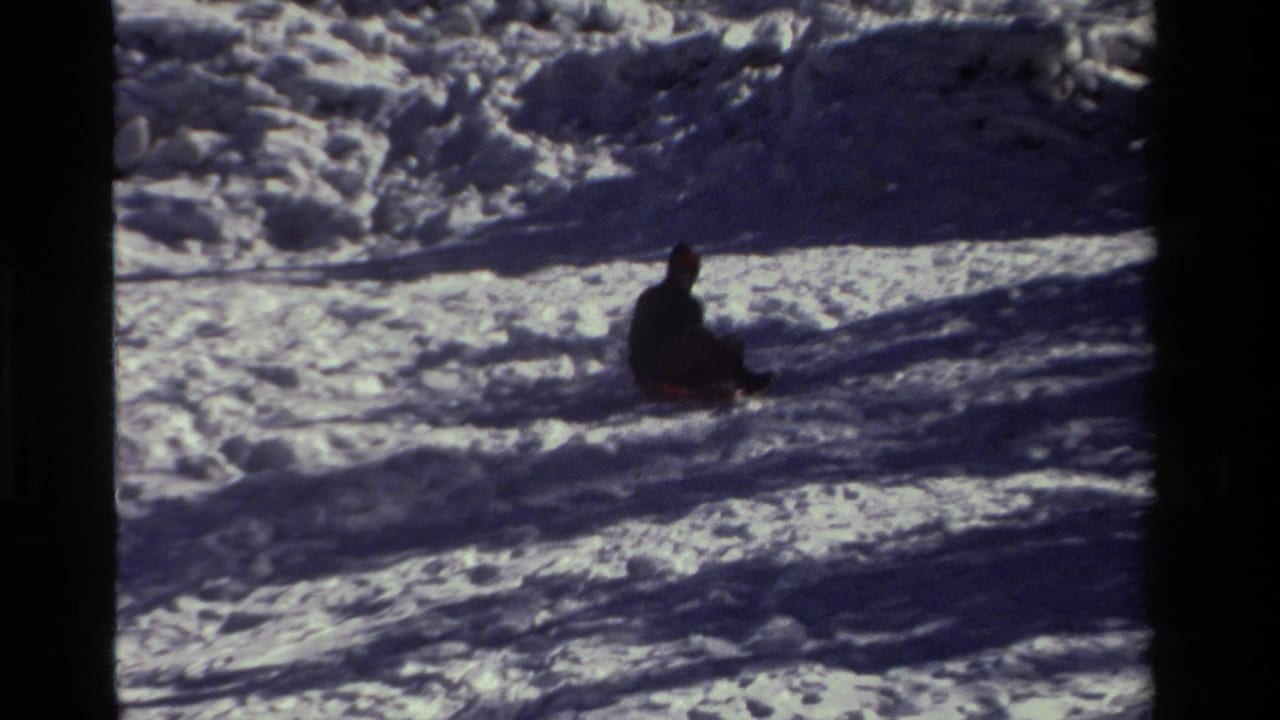 A bundled person sits on a snowy slope with long shadows across the snow.