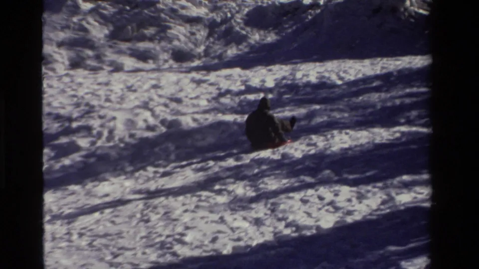 The 8mm frame shows the sledder farther downhill, seated low against textured snow and winter shadows.