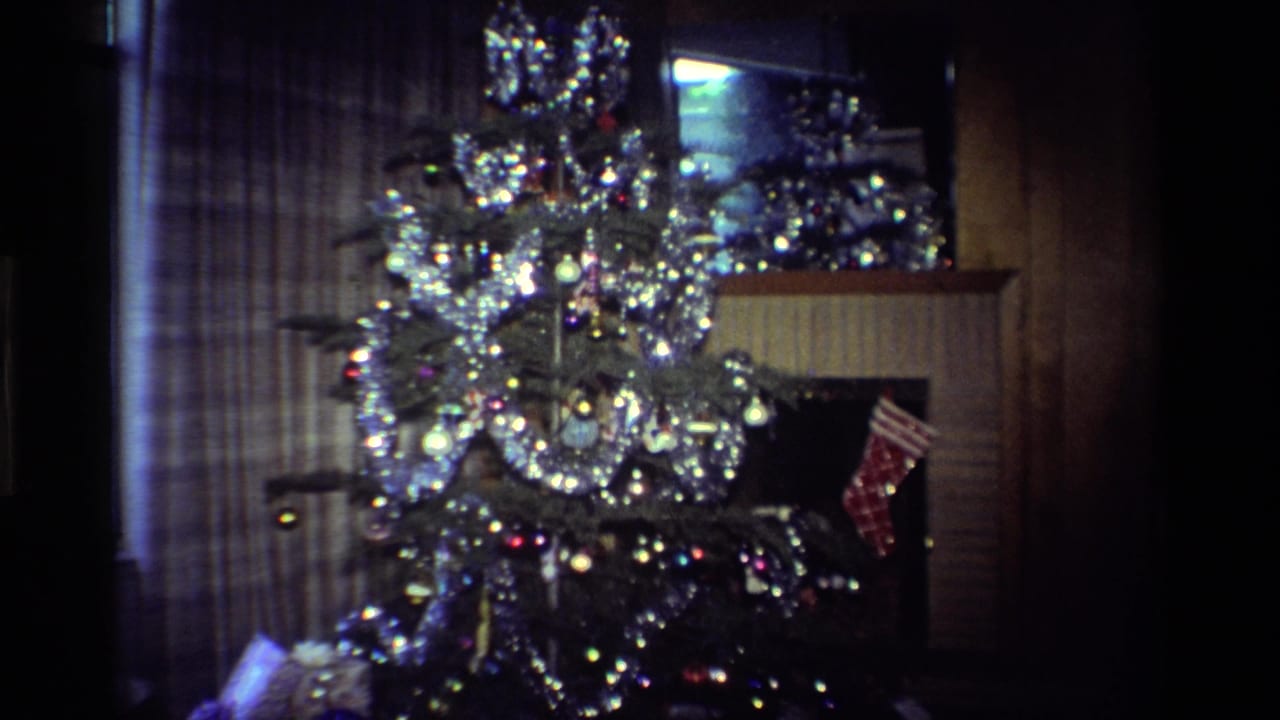 1984 California home-movie frame showing a decorated Christmas tree with silver tinsel, ornaments, wrapped gifts, and a stocking by the fireplace.