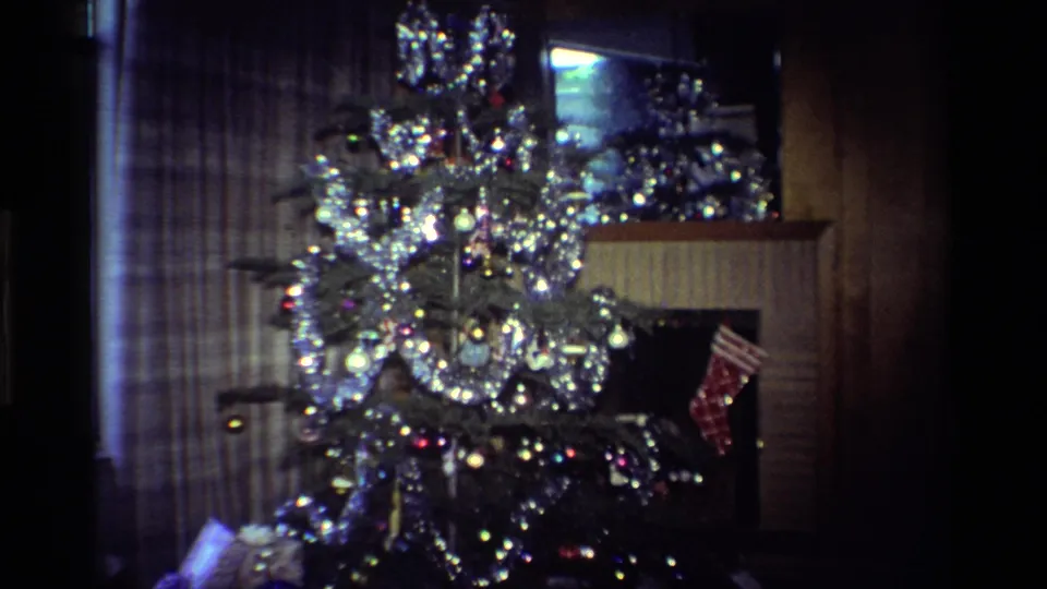 1984 California home-movie frame showing a decorated Christmas tree with silver tinsel, ornaments, wrapped gifts, and a stocking by the fireplace.