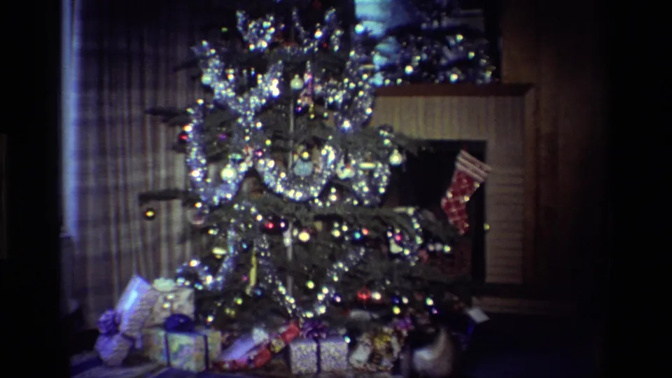 1984 home-movie frame of a decorated Christmas tree with reflective tinsel, baubles, wrapped presents, curtains, and fireplace details.