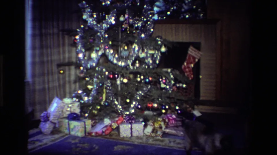 California 1984 home-movie frame showing the decorated tree, wrapped presents, fireplace stocking, and a cat partly visible near the floor.