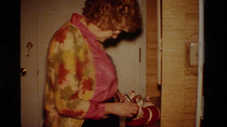 A woman in a bright patterned jacket stands in a private California interior near a doorway in 1984 home-movie footage.