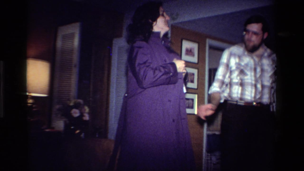 In a 1984 California home interior, a woman in a purple coat stands near a bearded man in glasses as he looks toward her arm.
