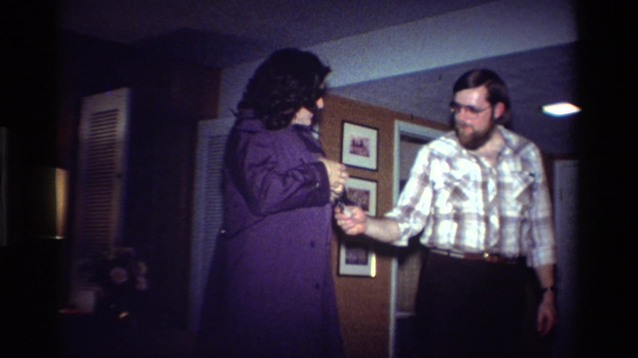 In a 1984 California home setting, a man in glasses gestures toward a woman in a purple coat with an arm-worn badge.