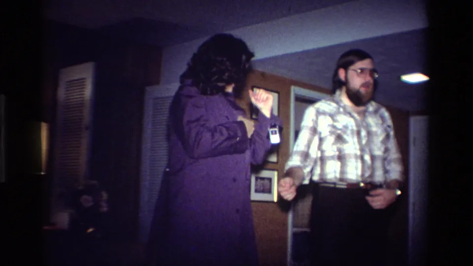 A woman in a purple coat raises her hand near an ID badge as a bearded man in glasses watches inside a 1984 California home.