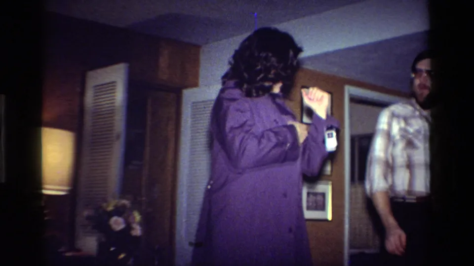 In the final 1984 California interior frame, the woman remains by the badge while the man in plaid moves toward the edge of the shot.