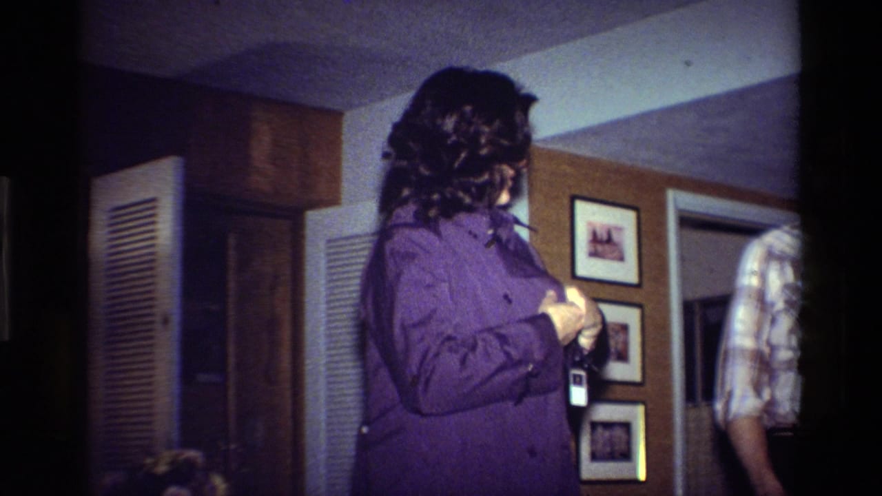 In a 1984 California home-movie frame, a dark-haired woman adjusts a purple shirt beside framed photos and louvered doors.