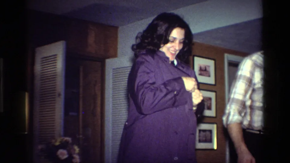 The woman smiles while adjusting a purple shirt in a 1984 California private home, with a plaid-shirted man partly visible.