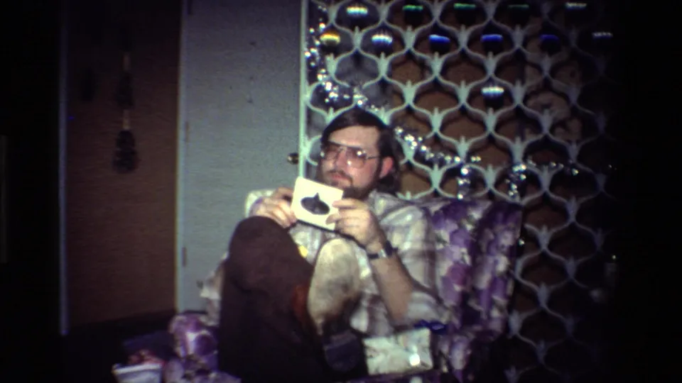 A bearded man with large glasses sits in a patterned chair holding a small card inside a 1984 California home.