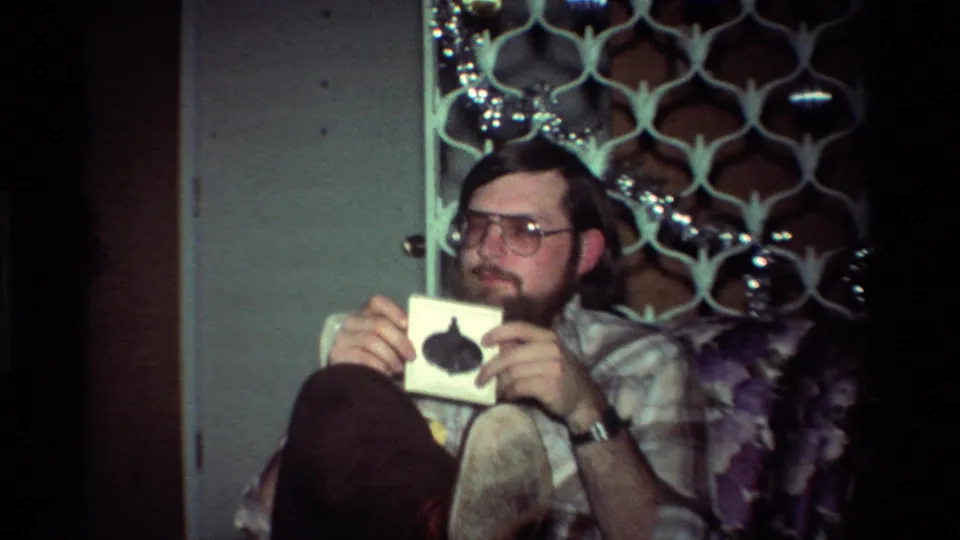 The seated man holds a small card near his chest in a dim 1984 California home interior with patterned furniture behind him.