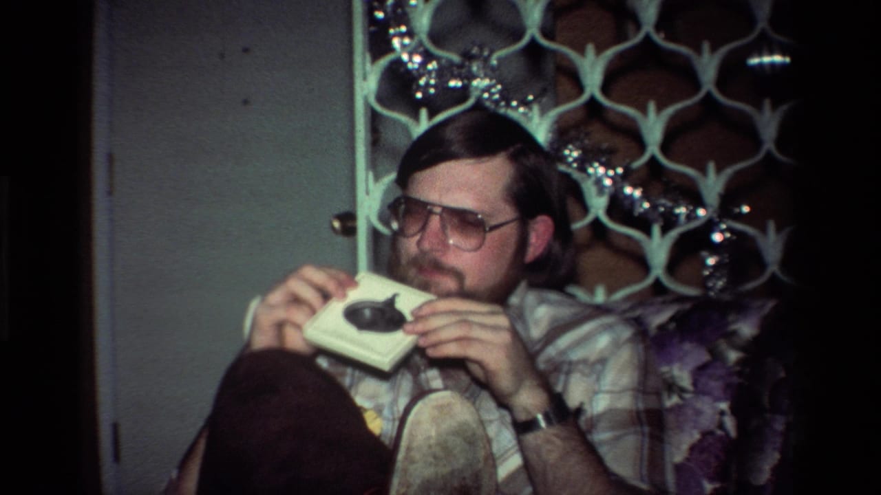 Man in glasses sits in a chair holding a small white object near his face.