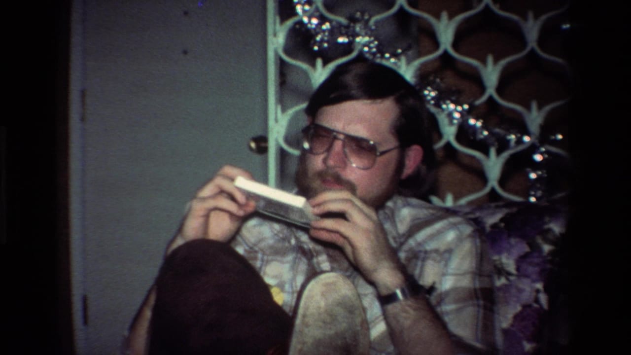 The man remains seated in the California interior, holding the white object sideways as tinsel and a patterned screen appear behind him.