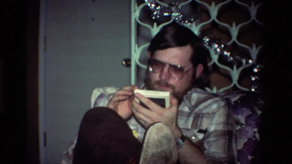 A close home-movie view shows the seated man looking down at a small white item with a dark circular graphic or opening.