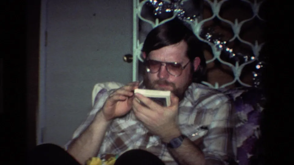 The man continues examining the small white object in a dim California private interior with tinsel and a decorative screen behind him.
