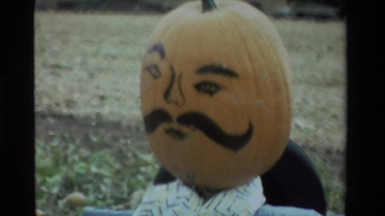 Close 8mm view of a painted pumpkin-headed scarecrow in a farm field, with eyebrows, eyes, nose, and mustache visible.