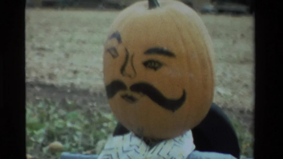 Close 8mm view of a painted pumpkin-headed scarecrow in a farm field, with eyebrows, eyes, nose, and mustache visible.
