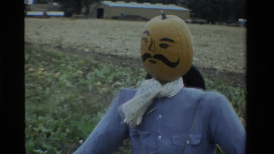 1984 California home-movie frame of a pumpkin-headed scarecrow in a blue shirt and patterned scarf, standing in a farm field.